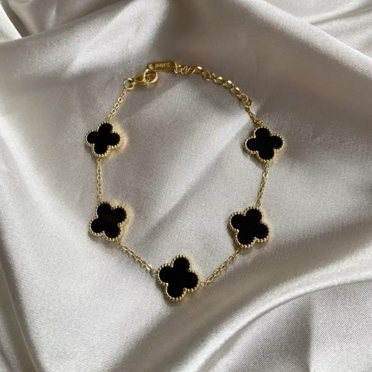 A gold chain bracelet with five black four-petal clover motifs, styled on a draped white satin fabric background.