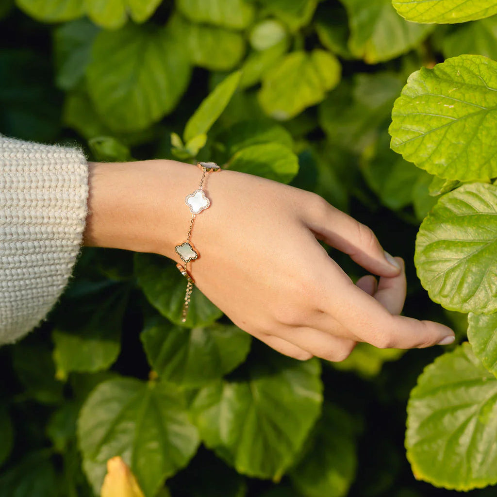A delicate gold-tone bracelet with a white clover charm and a white rectangular stone accent, worn on a person's wrist against a background of lush green leaves.